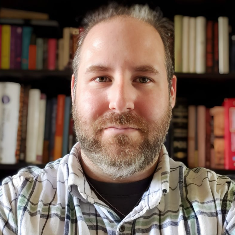 A man with beard and plaid shirt in front of bookshelves.